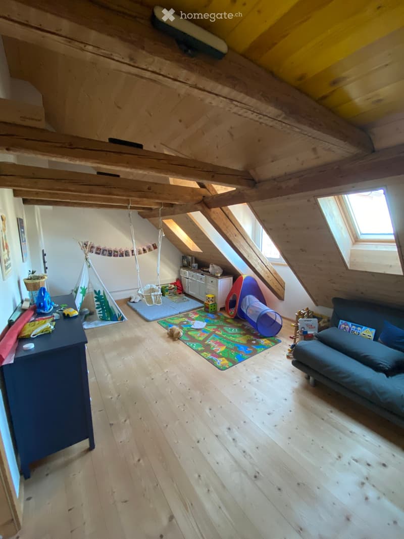 A cozy attic room with wooden floors and slanted ceilings, featuring a sofa, play area with a tent, swing, and various toys.
