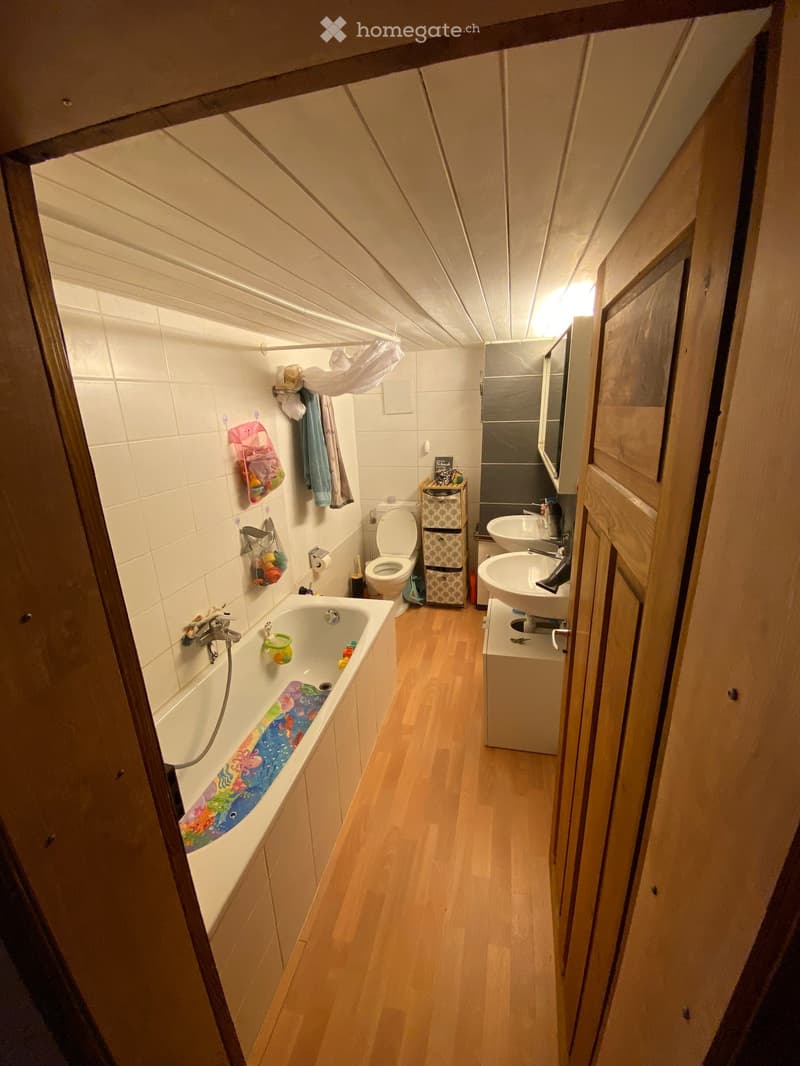Bathroom with a bathtub, toilet, double sink, mirror, and storage unit. Wooden flooring and white tiled walls.