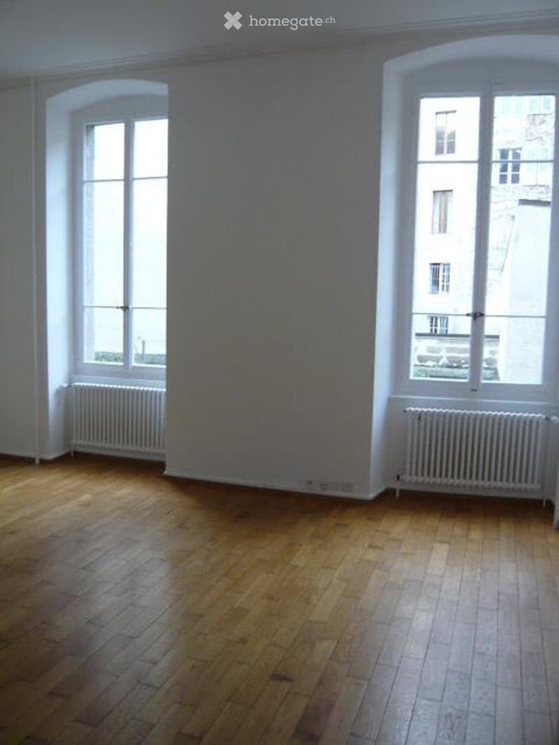 Empty room, hardwood floor, white walls, two large windows, white radiators, natural light from windows.