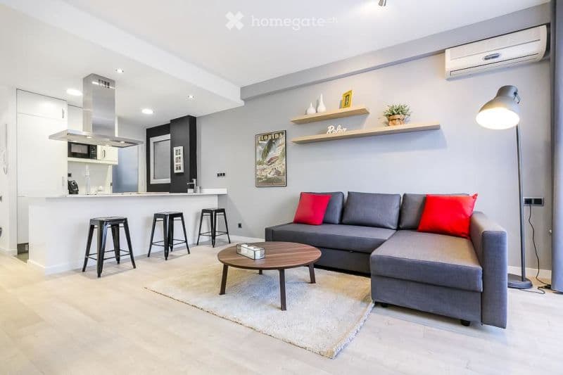 Modern open-concept living and dining area with grey sectional sofa, red throw pillows, wooden coffee table, bar stools, and a small kitchen area with a microwave and island.