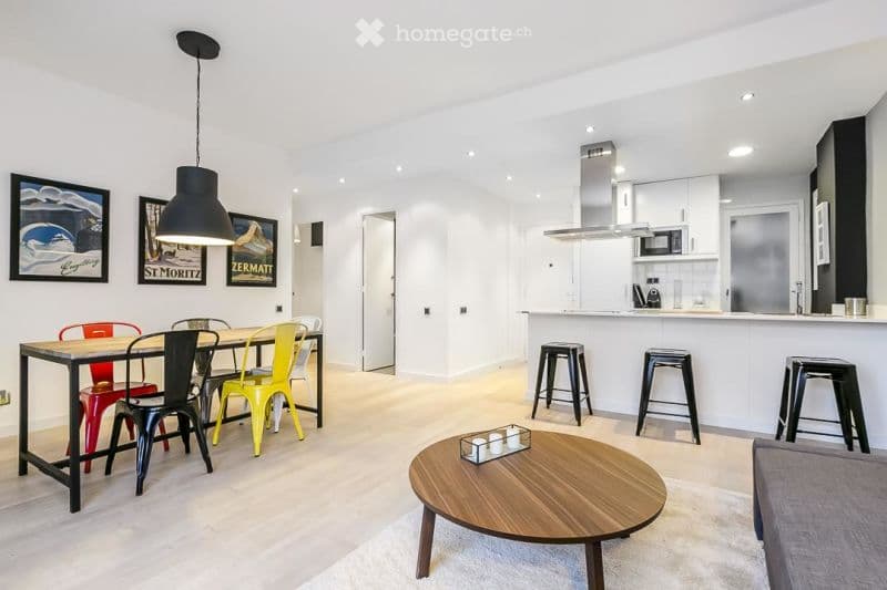 Open-plan living and dining area with modern furniture, including a dining table with colorful chairs, a coffee table, a sofa, and kitchen island with stools. The space features white walls, light wood flooring, and contemporary decor.