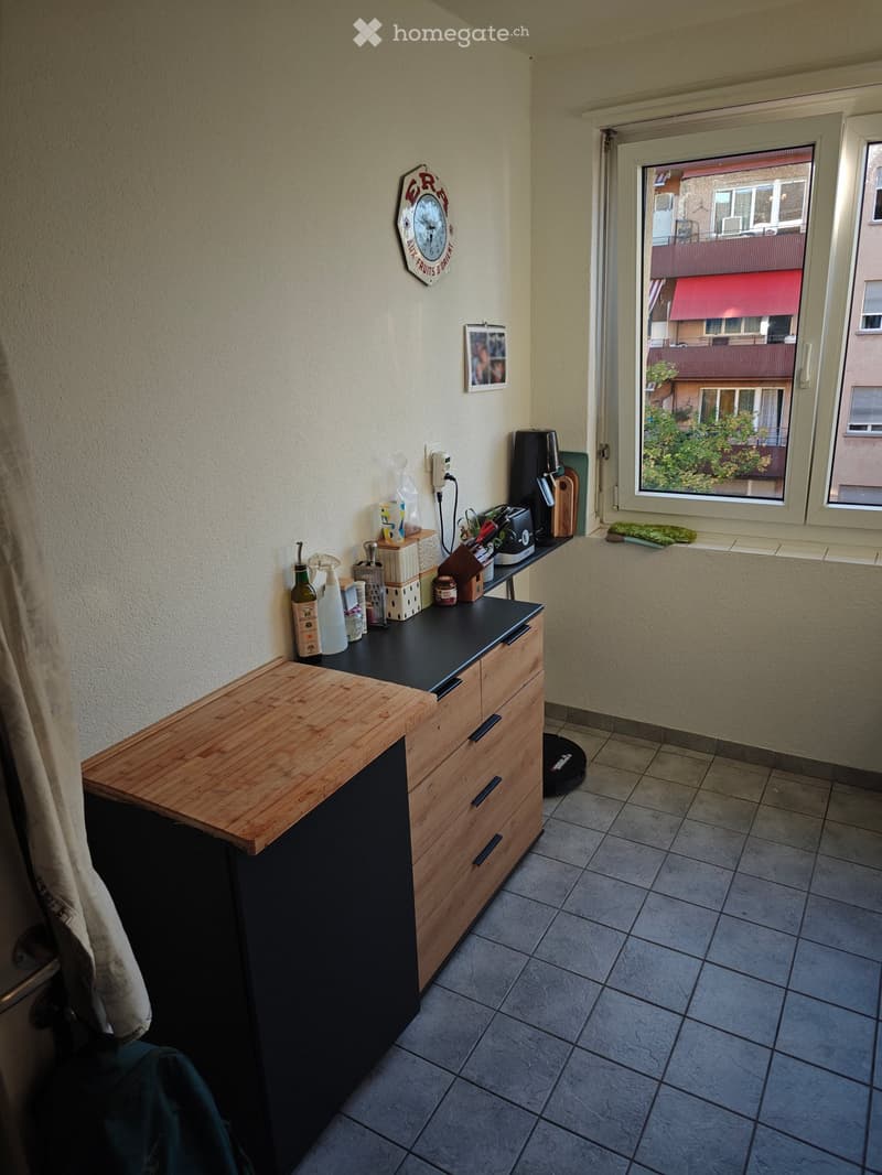 Compact kitchen with wooden counter, black countertop, drawers, and various small appliances and items on the counter. A window offers a view of the street.