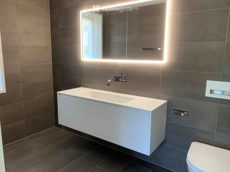 Modern bathroom with a white sink, chrome faucet, large illuminated mirror, and a toilet. Dark tiled walls and floor.