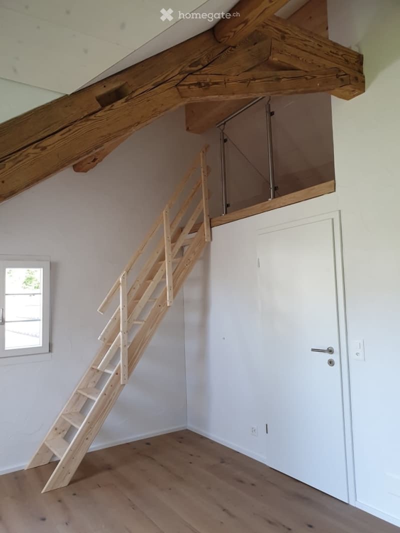 An attic space with wooden beams, a wooden staircase leading to a loft area, white walls, and a small window.