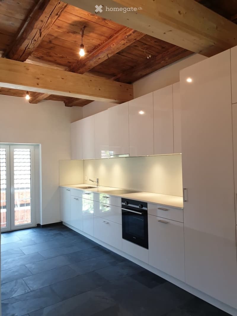 Modern kitchen with white cabinets, stainless steel sink, and built-in oven. Exposed wooden beams on the ceiling and large windows provide natural light.