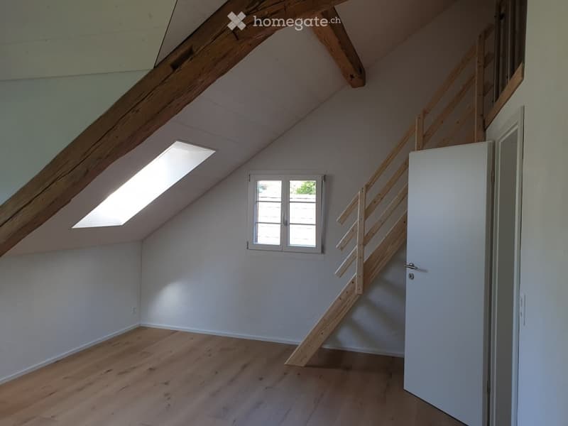 Empty attic room with wooden floors, white walls, skylight window, and wooden staircase leading up.