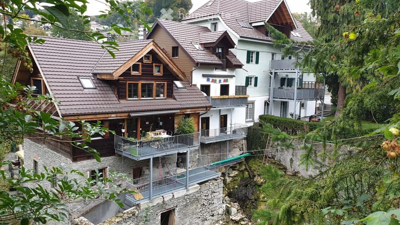 A cluster of multi-story houses with brown tiled roofs, wooden facades, and balconies, nestled in a lush, green hillside setting.