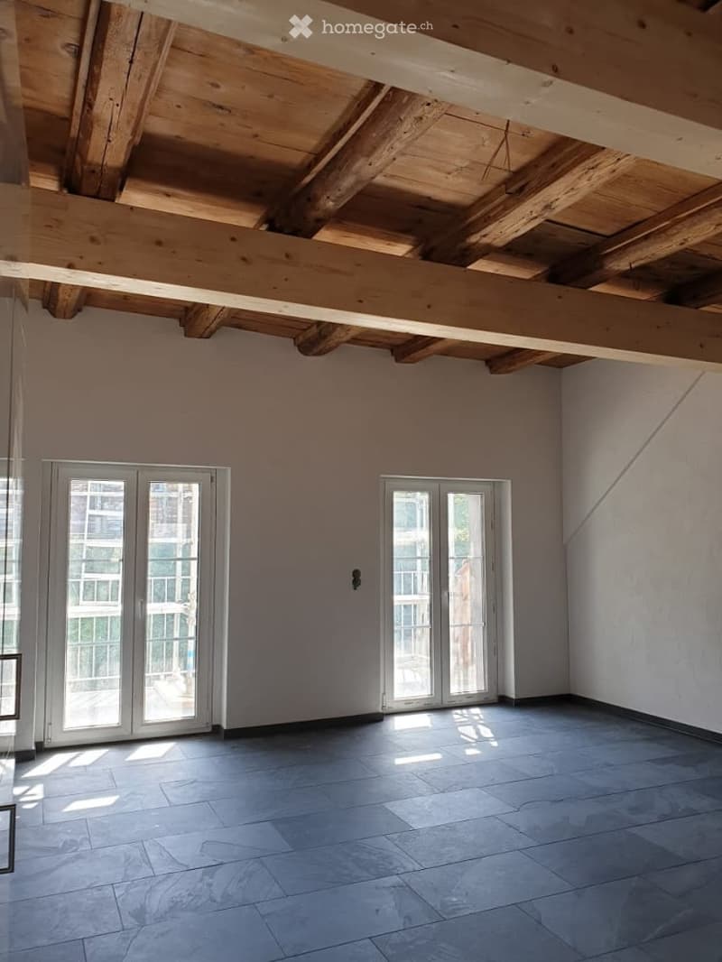 Empty room with exposed wooden ceiling beams, large windows, and tiled floor.