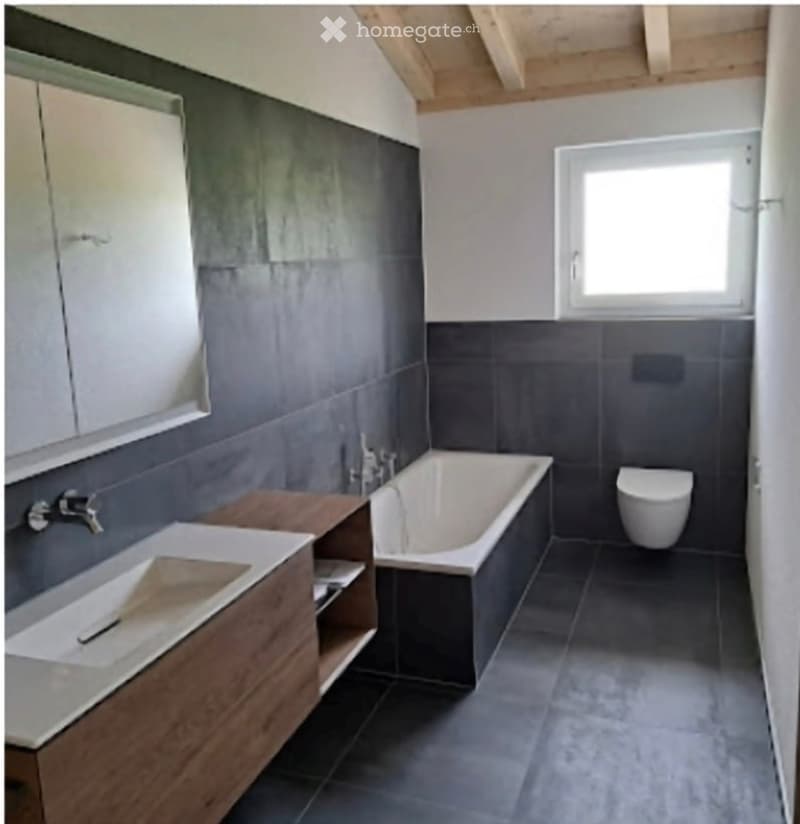 Modern bathroom with dark grey tiles, a wooden vanity with a sink, a bathtub, and a toilet. The room features a large window and a sleek, contemporary design.