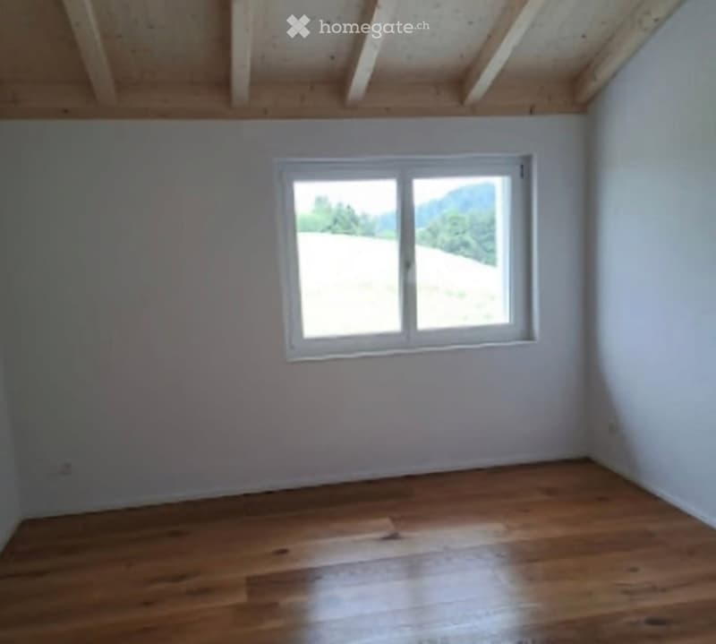 Empty room with wooden flooring, white walls, and a single window offering a view of a green landscape.