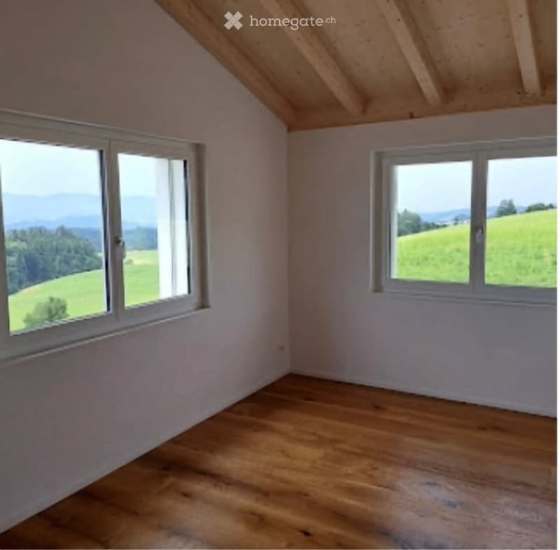 Empty room with wooden floor, white walls, and large windows offering scenic views of green fields and mountains.