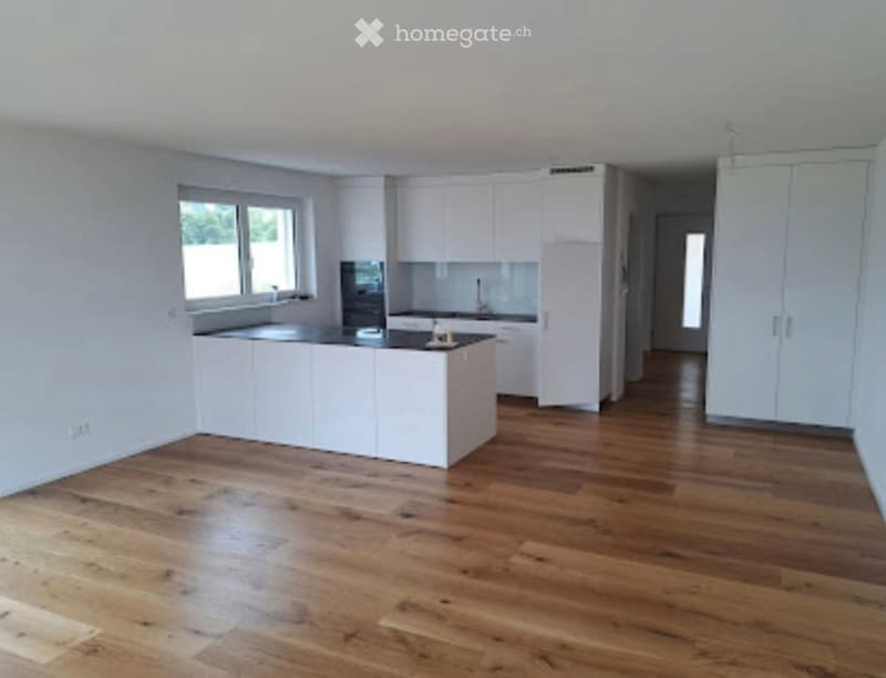 Empty kitchen with white cabinets, wooden flooring, and a large window letting in natural light. No appliances or furniture are present.