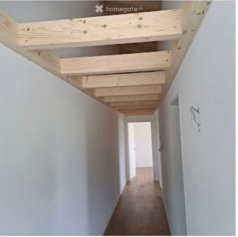 Empty hallway with exposed wooden beams on the ceiling, white walls, and a wooden floor.