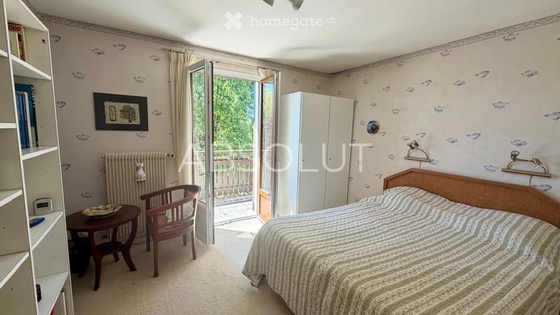Cozy bedroom with a bed, chair, small table, radiator, wardrobe, shelves, and a picture frame. The room has floral wallpaper and opens to a balcony.