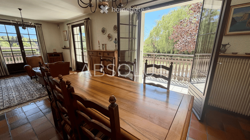 Bright dining room with large windows offering a scenic view, wooden dining table with chairs, leather sofa, armchair, cabinet, chandelier, and decorative picture frames.