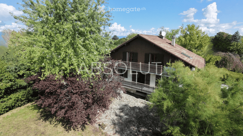 A two-story house with a brown roof and white balcony, surrounded by lush greenery and trees, offering a scenic view.