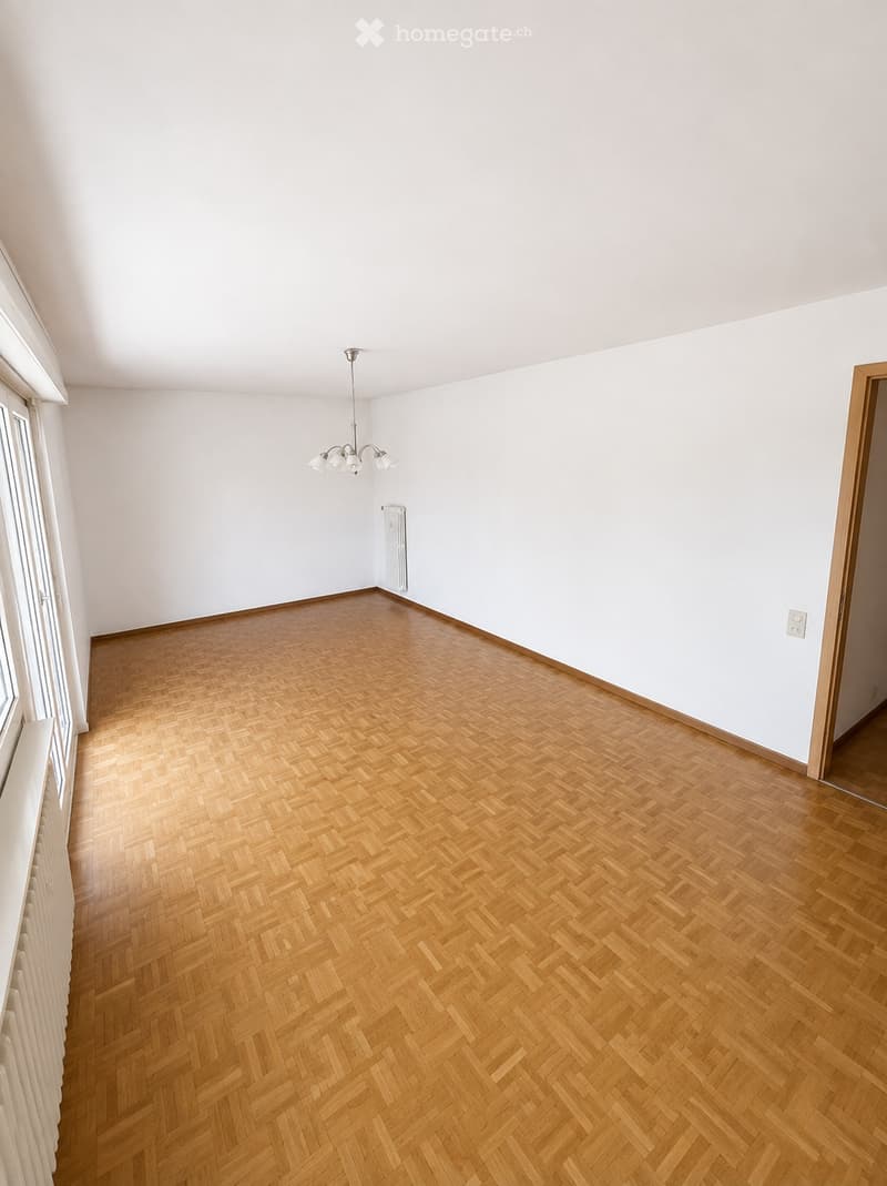 Empty room with wooden herringbone flooring, white walls, and a ceiling light fixture. The room has large windows on one side and a radiator along the wall.