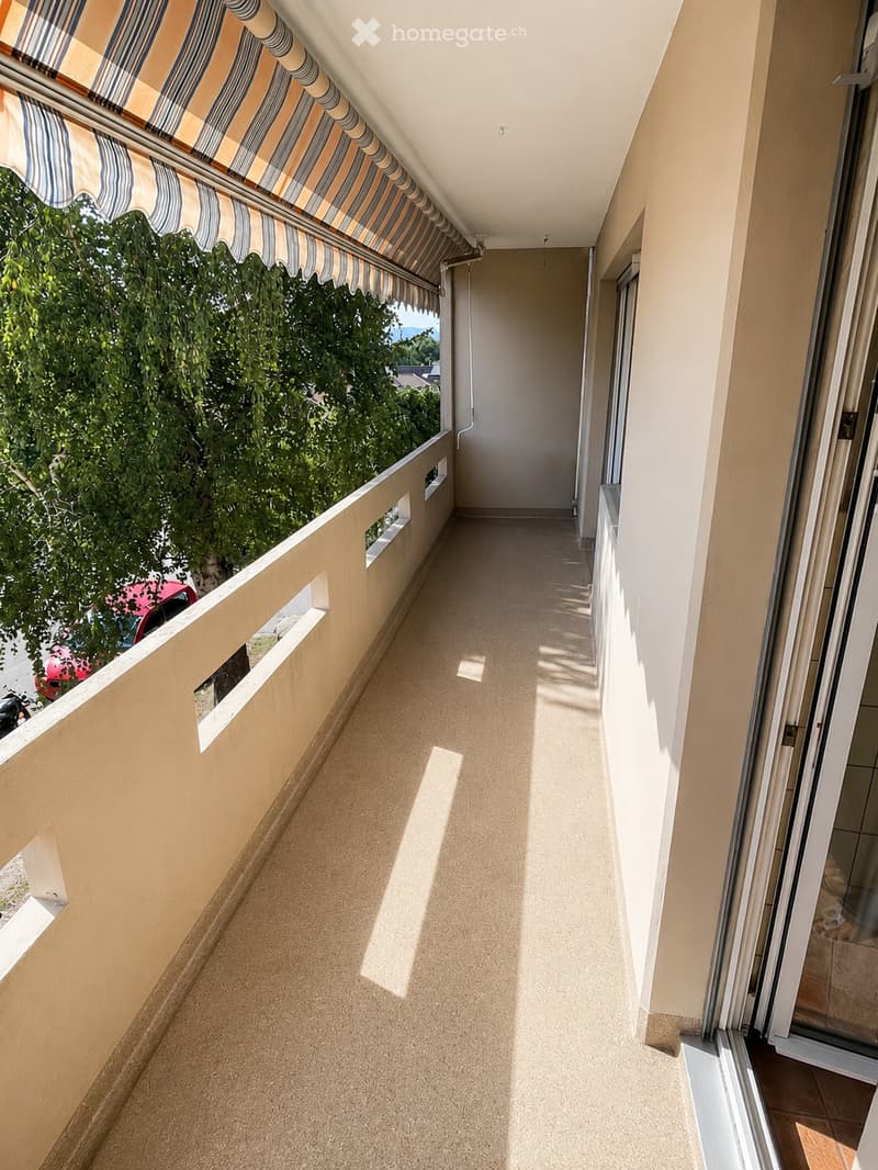 Balcony with striped awning, beige walls, and tiled floor, overlooking greenery.