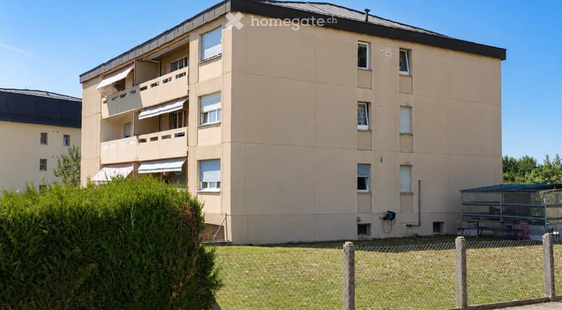 Four-story residential building with beige facade, multiple balconies, and a fenced yard with outdoor seating area.