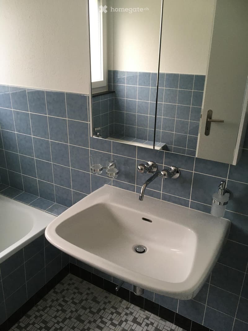 Bathroom with a white sink, chrome faucet, mirror, two cups, and a soap dispenser. Blue tiled walls and a small window.