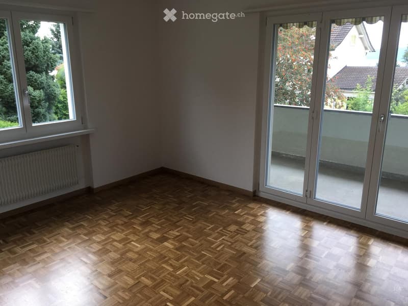 Empty room with wooden herringbone flooring, large windows, and sliding glass doors leading to a balcony.