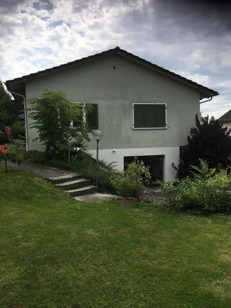 Single-story house with grey exterior, white trim, and a small porch. The house has a well-maintained lawn, garden, and steps leading to the entrance.
