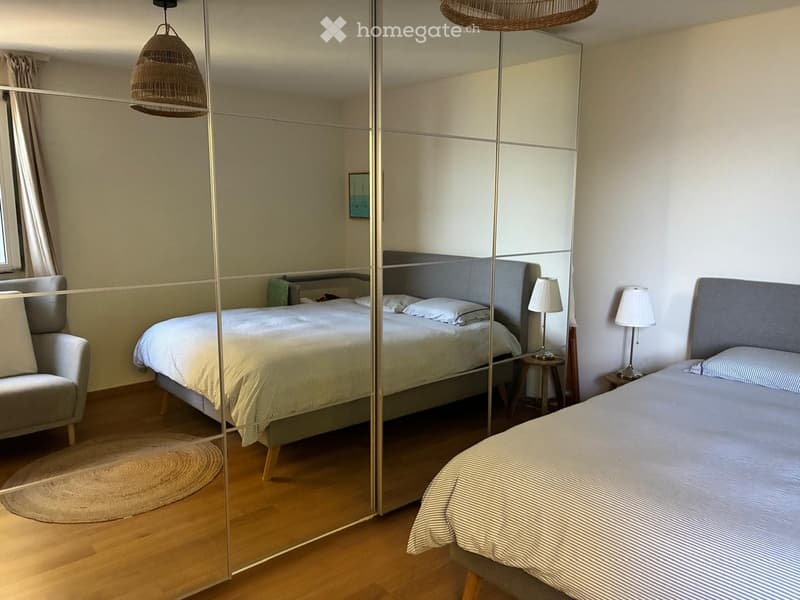Modern bedroom with a large mirrored wardrobe, a neatly made double bed, two bedside tables with lamps, a wicker pendant light, and a small armchair.