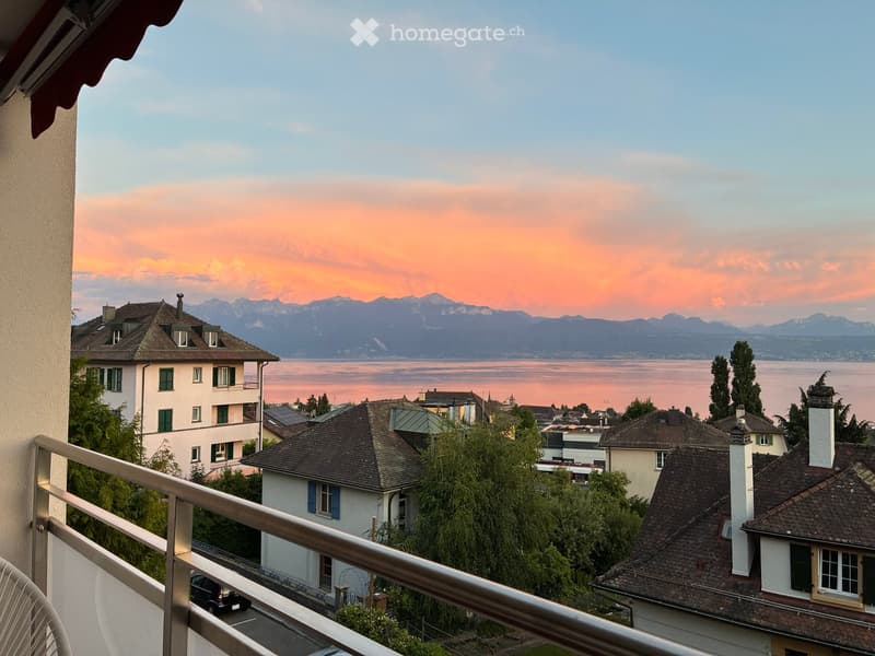 Balcony overlooking a scenic view of a lake and distant mountains at sunset, with several houses and trees in the foreground.