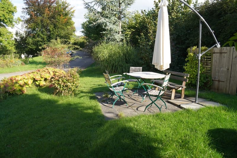 Garden area with a patio seating set including a table, four chairs, and a bench, surrounded by lush greenery and a large umbrella.