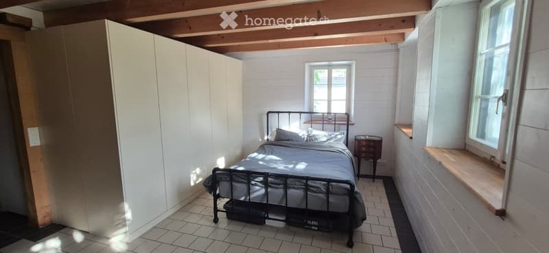 Cozy bedroom with a metal-framed bed, nightstand, and built-in wardrobe. The room features white shiplap walls, wooden beams on the ceiling, and a window with a built-in seat.
