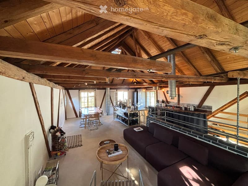 Open-plan living and dining area with exposed wooden beams, a dark-colored sofa, a small round coffee table, a dining table with chairs, and a modern kitchen area with a counter and appliances.