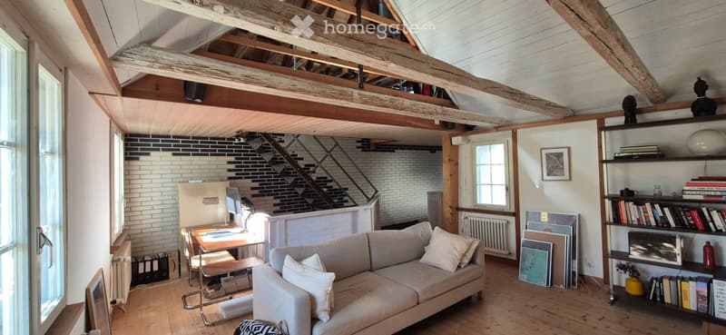 Loft-style living space with exposed wooden beams, a sofa, coffee table, bookshelf, desk, and artwork.