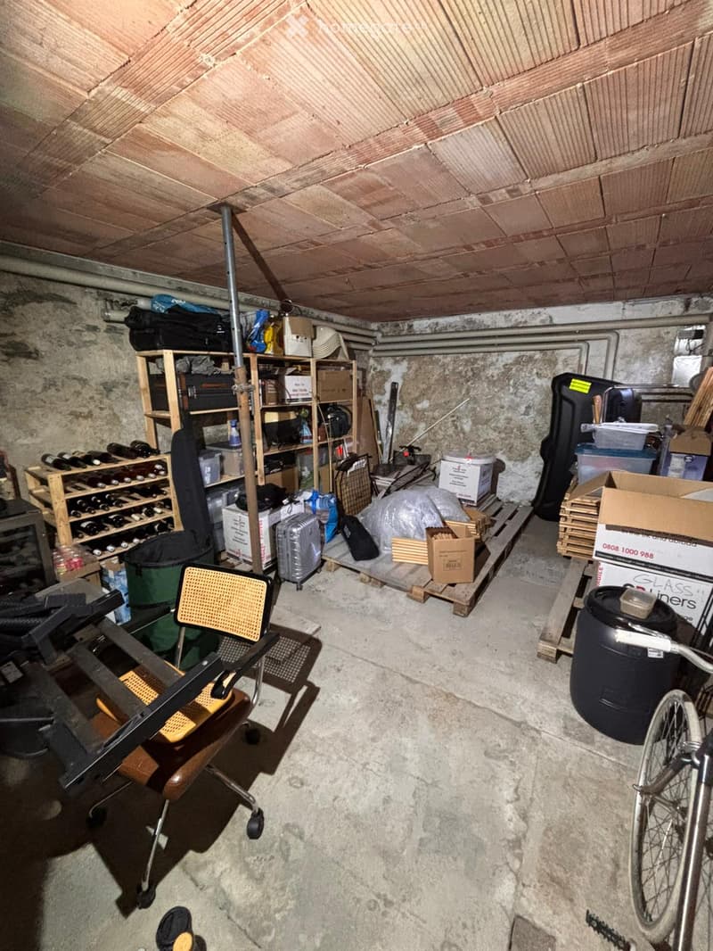 Storage room with concrete floor, exposed wooden ceiling, shelves holding wine bottles, suitcases, and storage boxes. A chair, bicycle, and barrel are also present.