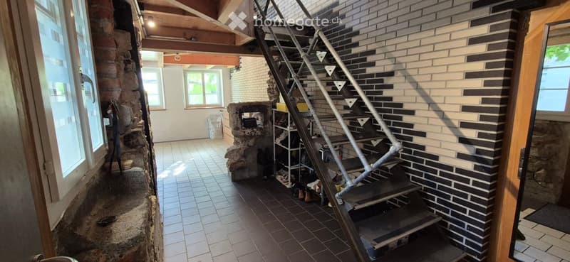 A rustic hallway with exposed wooden beams, a stone wall, and a metal staircase leading upwards. The floor is tiled, and there are windows allowing natural light to enter.