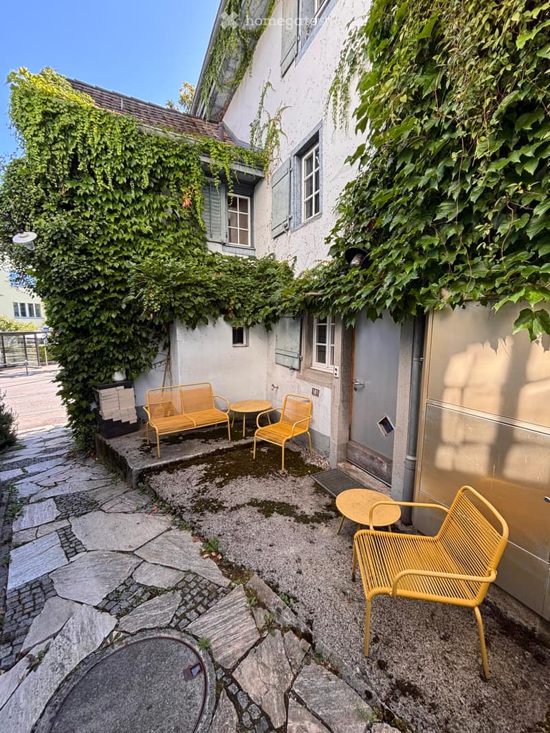 A small, cozy outdoor area with stone paving, yellow metal furniture including a bench, chairs, and a small table, surrounded by a building with ivy-covered walls.