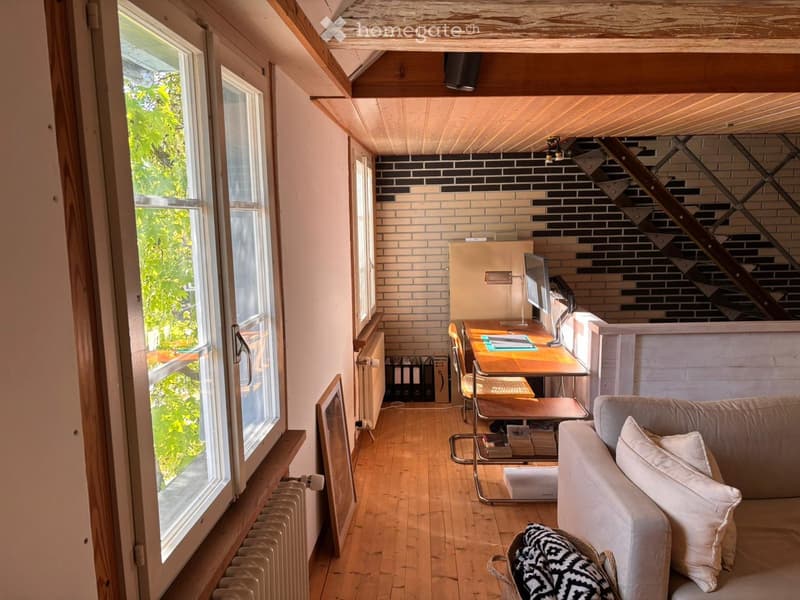 A cozy, multi-functional room with a slanted wooden ceiling, large windows, a sofa, a desk with a computer, and a staircase leading upwards.