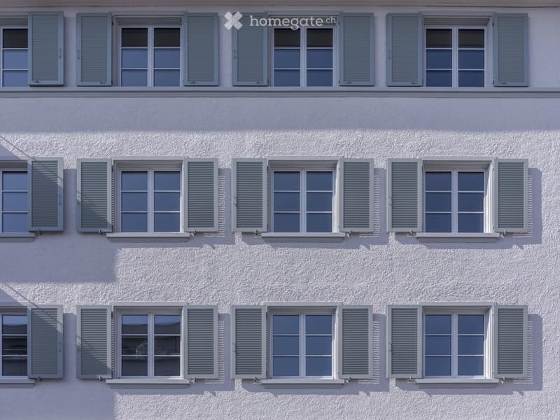 White building facade with multiple windows featuring grey shutters, arranged in a grid pattern across three visible floors.