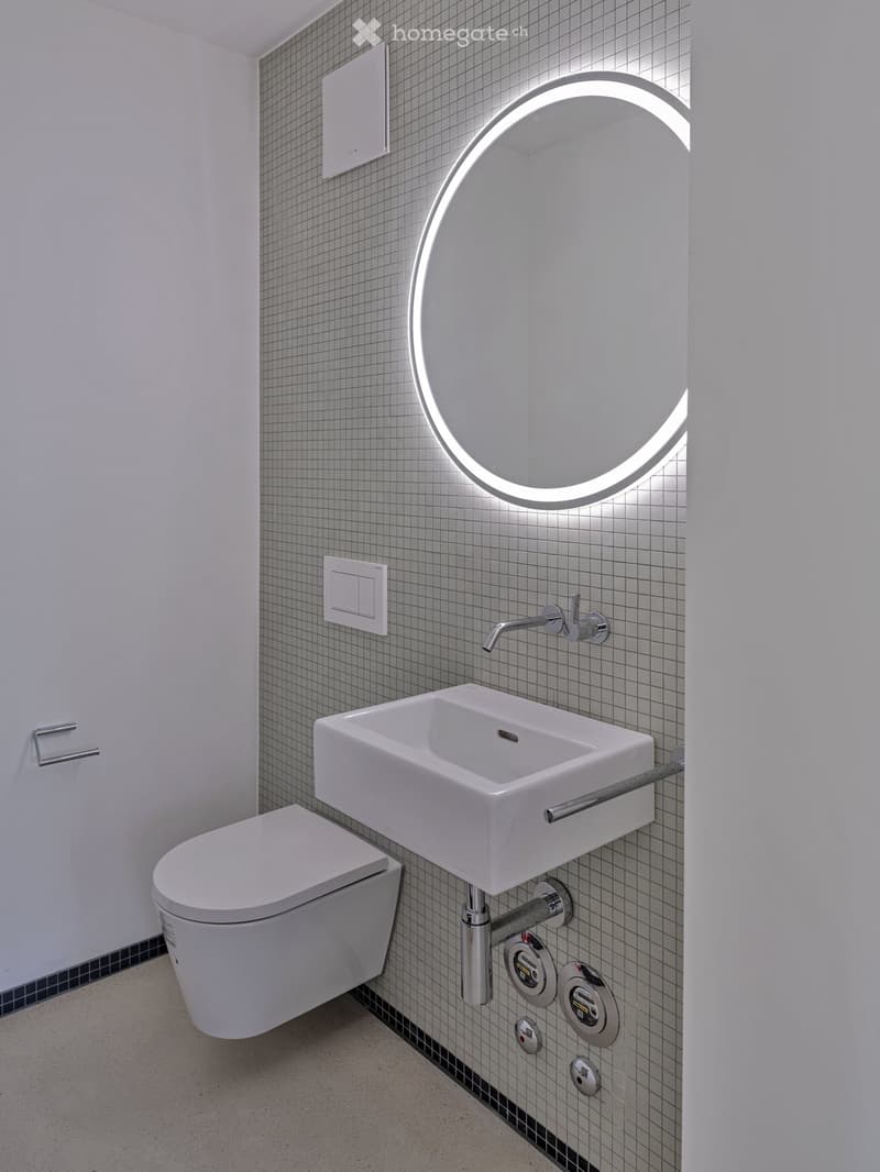 Modern bathroom with a white toilet, wall-mounted sink, chrome faucet, and illuminated round mirror.