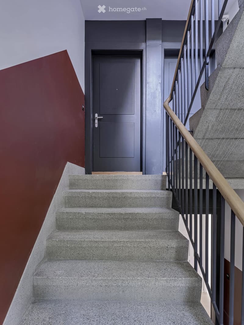 Modern staircase with concrete steps, wooden handrail, and metal balustrade, leading to a grey door.