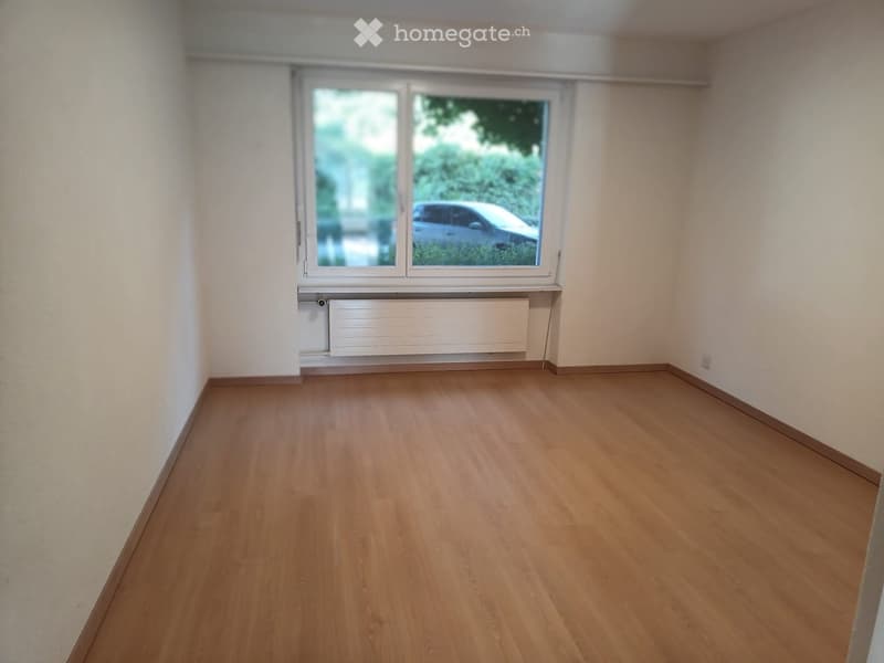 Empty room with wooden flooring, white walls, and a large window with a view of a car and greenery outside.