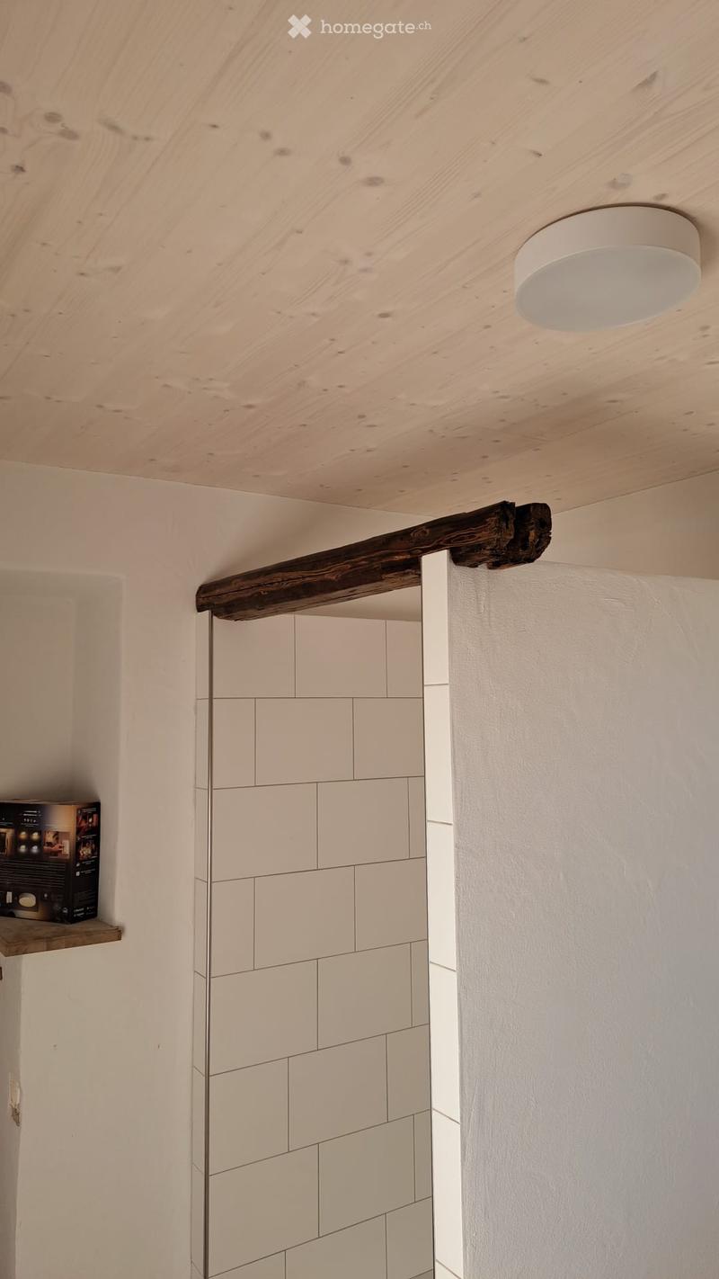 An empty room with a wooden beam on the ceiling and white tiled wall. There is a shelf with a box on it.