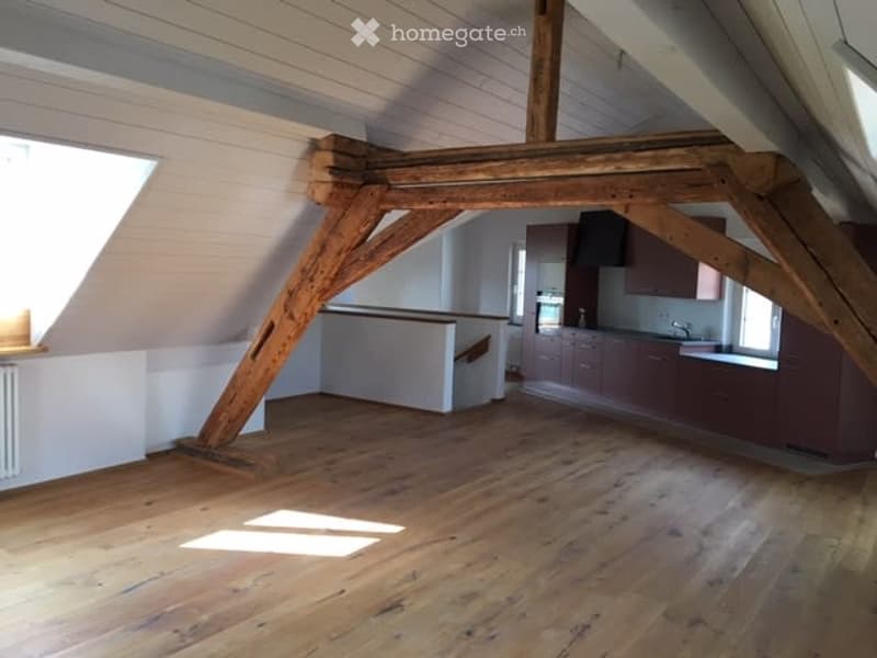 An attic space with wooden beams and exposed wooden floorboards, featuring a small kitchen area with cabinets and a sink, and a staircase leading to another level.