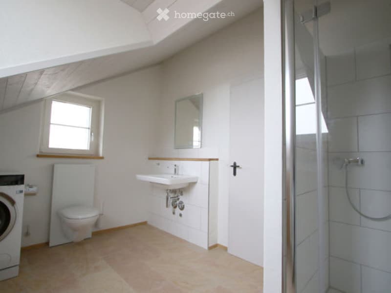 Bright, modern bathroom with a slanted ceiling, white walls, and light-colored tiles. Features include a sink with a mirror, a toilet, a shower, and a washing machine.