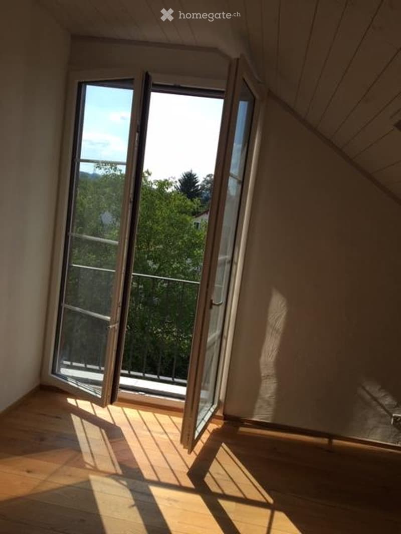 Empty room with wooden flooring, large open sliding doors leading to a balcony with a metal railing and a view of greenery.