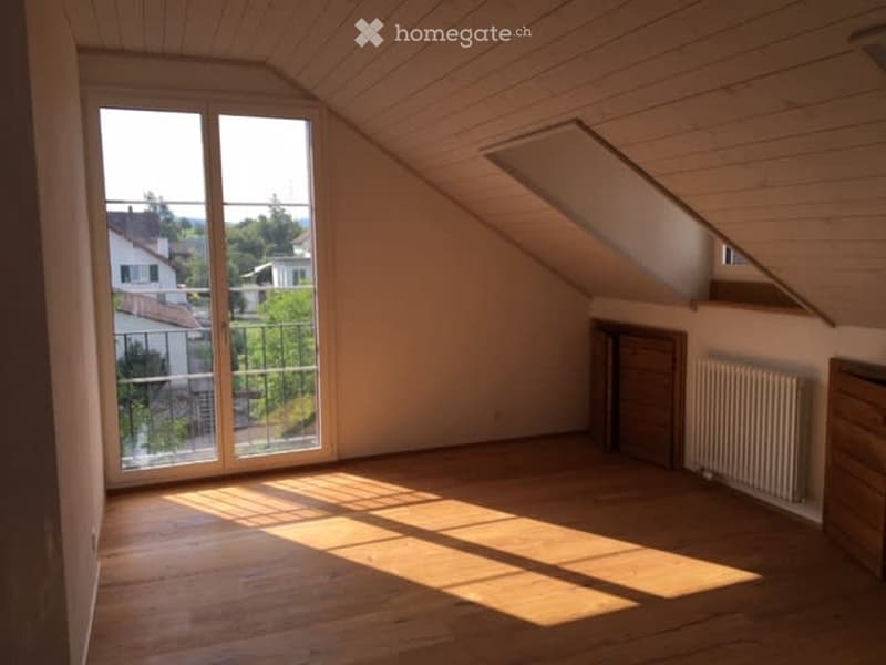 Bright, empty attic room with wooden floors, large windows, and a view of the surrounding area.