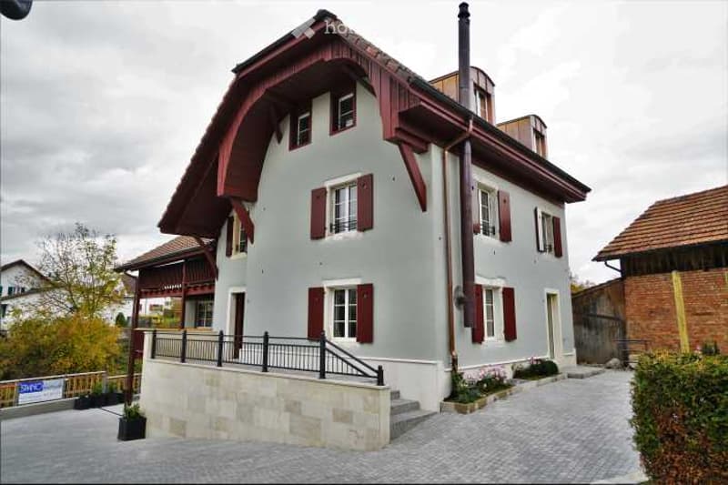 Two-story house with light blue walls, red shutters, and a raised ground floor with stone steps leading to the entrance.