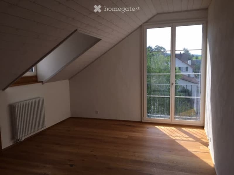 Empty attic room with wooden flooring, slanted ceiling, large window with glass door leading to a balcony, radiator on the wall, and natural light streaming in.