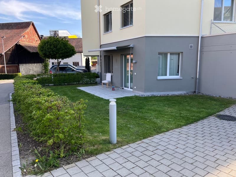 Well-maintained garden with a lawn, small trees, shrubs, and a paved pathway leading to the house entrance. A white chair is placed near the entrance, and a car is parked in the driveway.