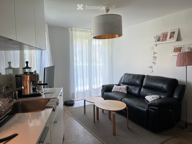 Modern open-plan living and kitchen area with a black leather sofa, wooden coffee table, small wooden stools, a kitchen counter with a coffee machine, and framed pictures on the wall.