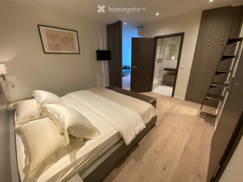 Modern bedroom with wooden floors, bed made with white sheets and pillows, walk-in closet, framed picture on the wall, modern decor, open door to a bathroom.